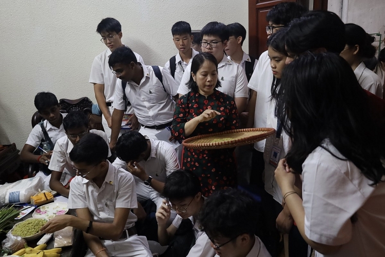 Cô giáo Lê Huyền, Phụ trách chương trình trao đổi du học sinh trường Raffles Institution và Trường THCS và THPT Nguyễn Tất Thành dẫn đoàn học sinh thăm và tìm hiểu Cốm Làng Vòng.  ອາຈານ ເລຮຽນ (Le Huyen), ຮັບຜິດຊອບໂຄງການແລກປ່ຽນນັກຮຽນຕ່າງປະເທດຢູ່ສະຖາບັນ Raffles ແລະ ໂຮງຮຽນມັດທະຍົມຕອນຕົ້ນ ຕອນປາຍ ຫງວຽນເຕີດແທງ, ໄດ້ນຳພາຄະນະນັກຮຽນໄປຢ້ຽມຊົມ ແລະ ຮຽນຮູ້ກ່ຽວກັບ Com Lang Vong.